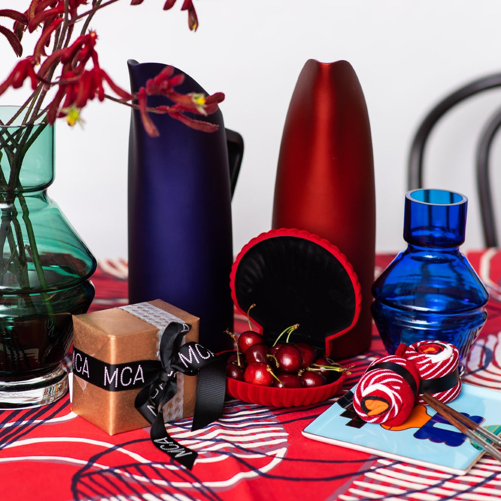 A scarlet red open clam shell containing red cherries sitting among a wrapped present, a green and blue glass vases and indigo and red FINK jugs on a red indigenous patterned tablecloth. 