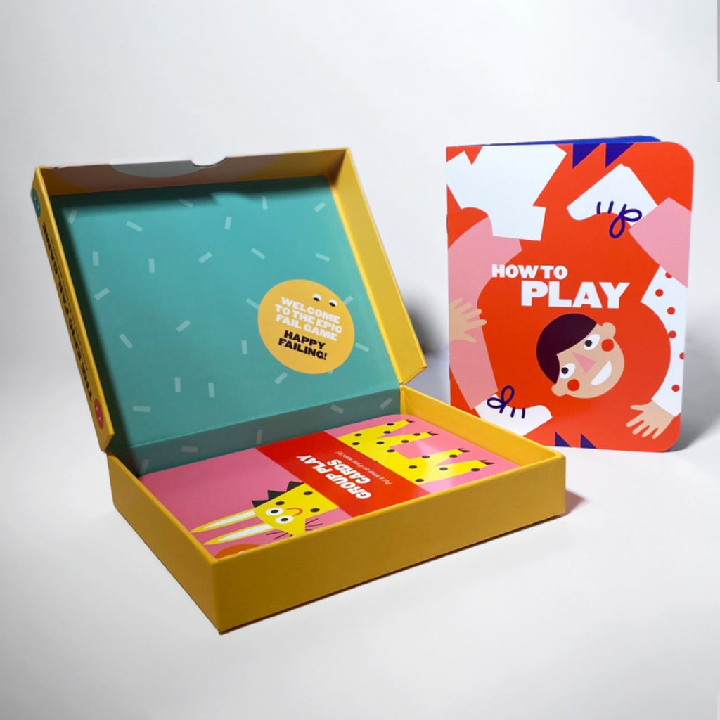 The opened yellow box reveals a teal interior with a yellow circle sticker "Welcome to the epic fail game. Happy failing!" and the bright coloured cards inside. Standing upright on the right is a red booklet with a quirky graphic of a body mixed up. 