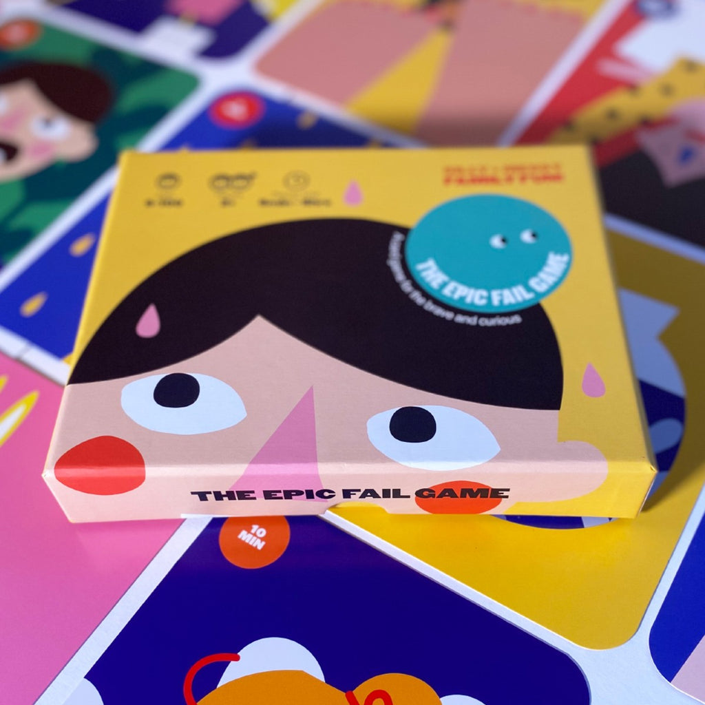 Sitting on top of a table laid with colourful cards in a grid is a yellow box illustrated with a face looking up and a teal circle on the brown hair with "the epic fail game" in white. 
