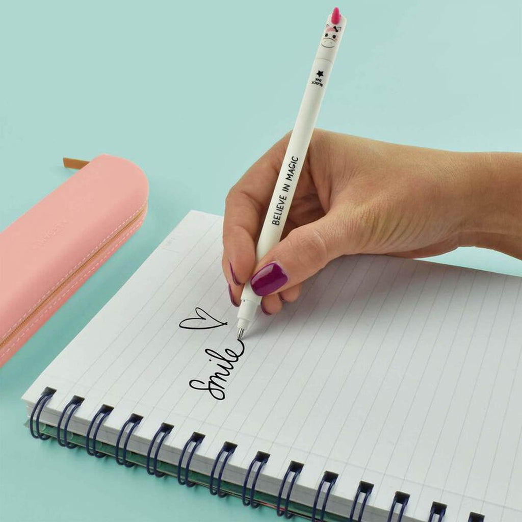 A hand writing on a lined notebook with a slender white pen with a unicorn face and a pink horn on the cap on a turquoise desk.
