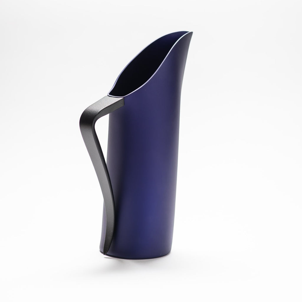 A indigo blue matte jug with its slender black handle facing towards the front, exposing the sculptural opening of the jug.  