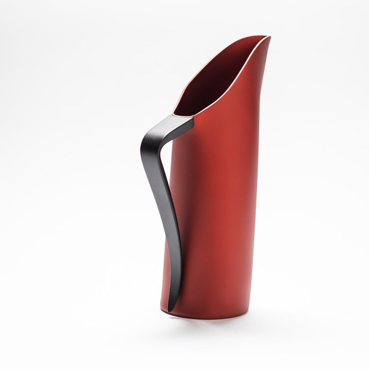 A red matte jug with its slender black handle facing towards the front, exposing the sculptural opening of the jug.