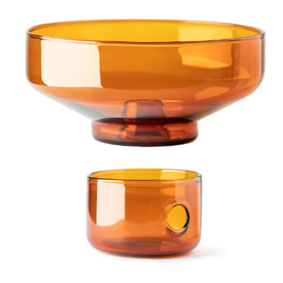 In front of the grey cube packaging box with the text “Oil & Water” on it is a amber glass oil burner. The top is conical shape that sits on top of short cylindrical glass with a round hole on the side.