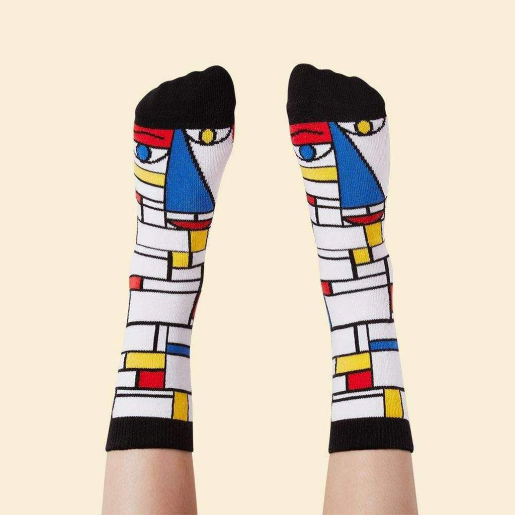 Against the pastel yellow background are a pair of feet wearing white socks in the pattern of Piet Mondrian's iconic 'Composition with Red, Blue, and Yellow' with his face in the same style by the toe end. 