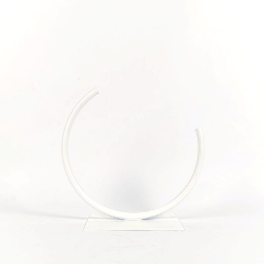 A white metal vase with a flat base has a thin pipe bent into an incomplete circle, forming a crescent shape. 