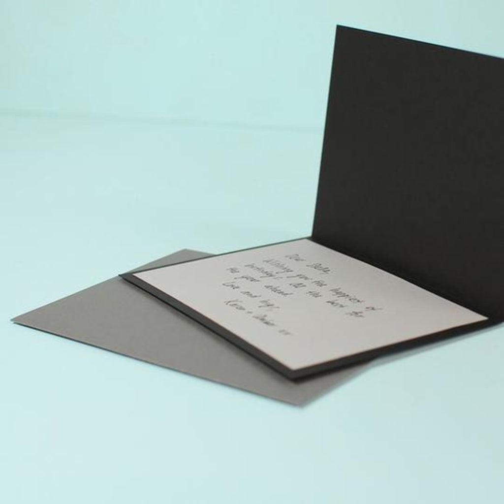 On an aquamarine background is an opened black greeting card with a handwritten message over a grey envelope. 