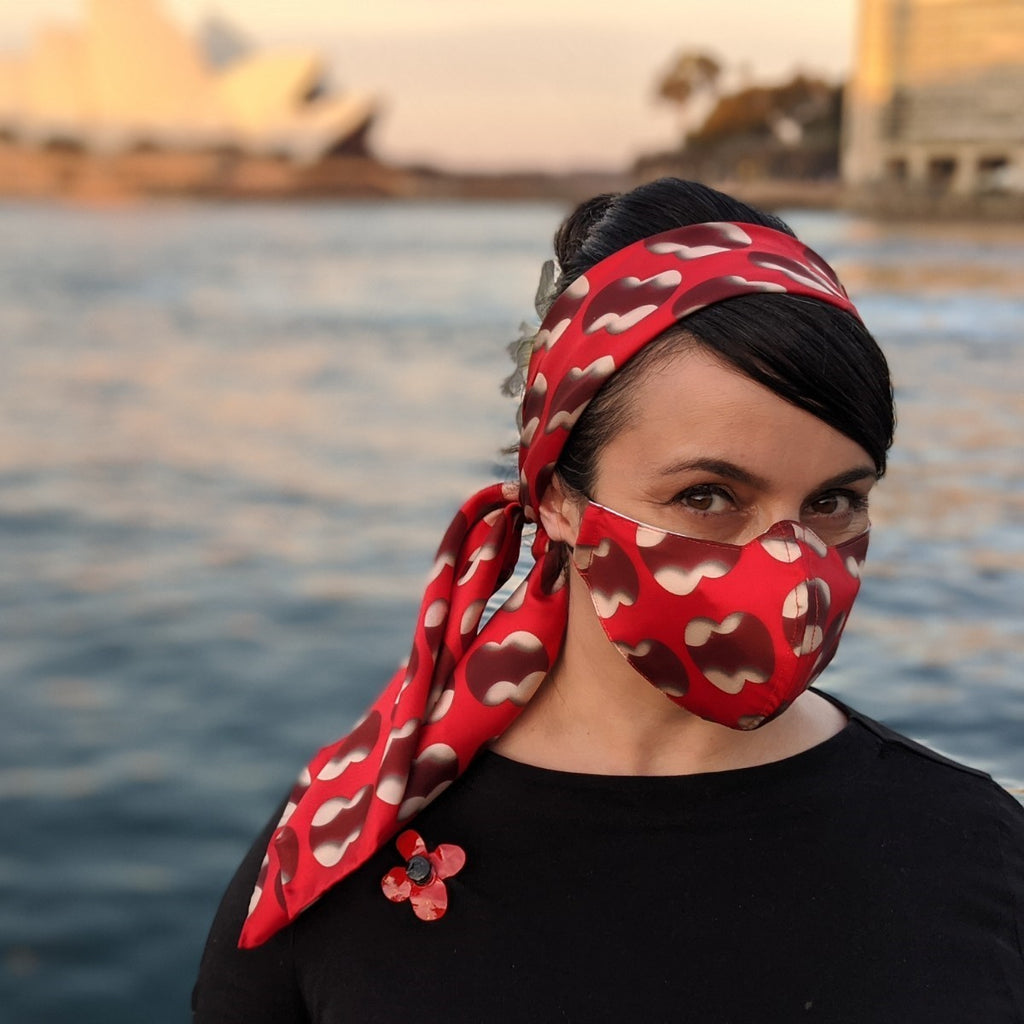 As the sun sets on the Sydney Opera House in the background, a woman wears a red face mask with a circular pattern and a matching headscarf.