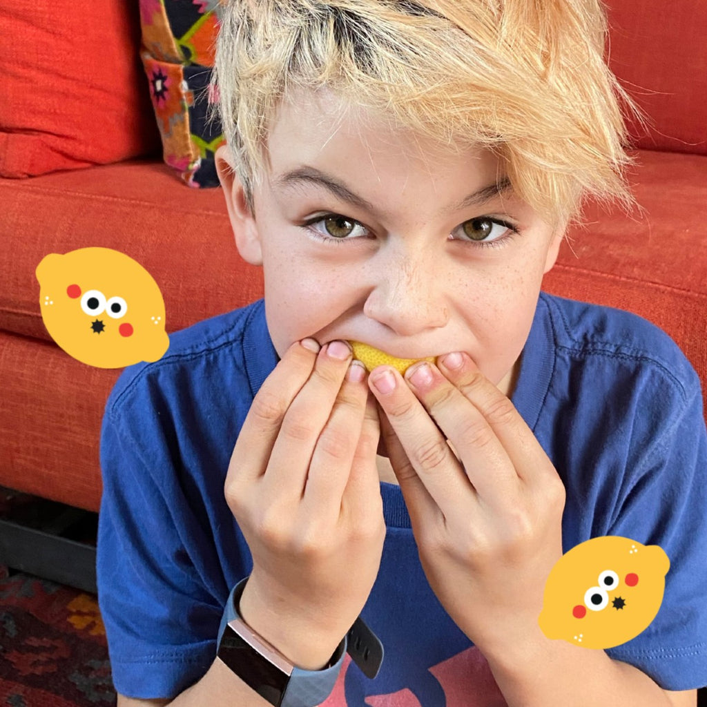 A blonde child is wearing a blue t-shirt eating a lemon in front of an orange sofa. On both sides of the child's shoulder are two lemon graphic stickers making a sour face.