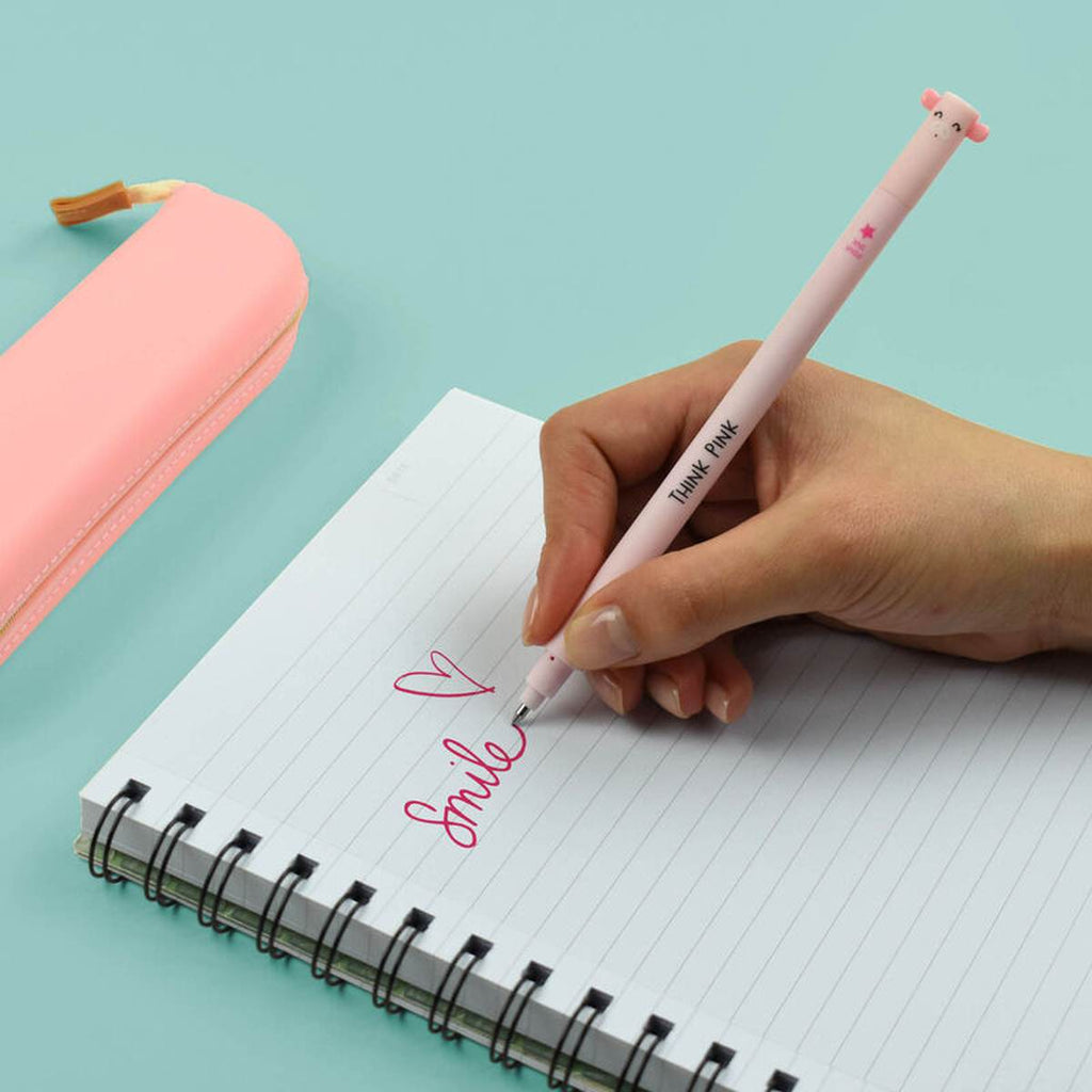 A hand writing on a lined notebook with a slender pink pen with a pig face on the cap on a turquoise desk.