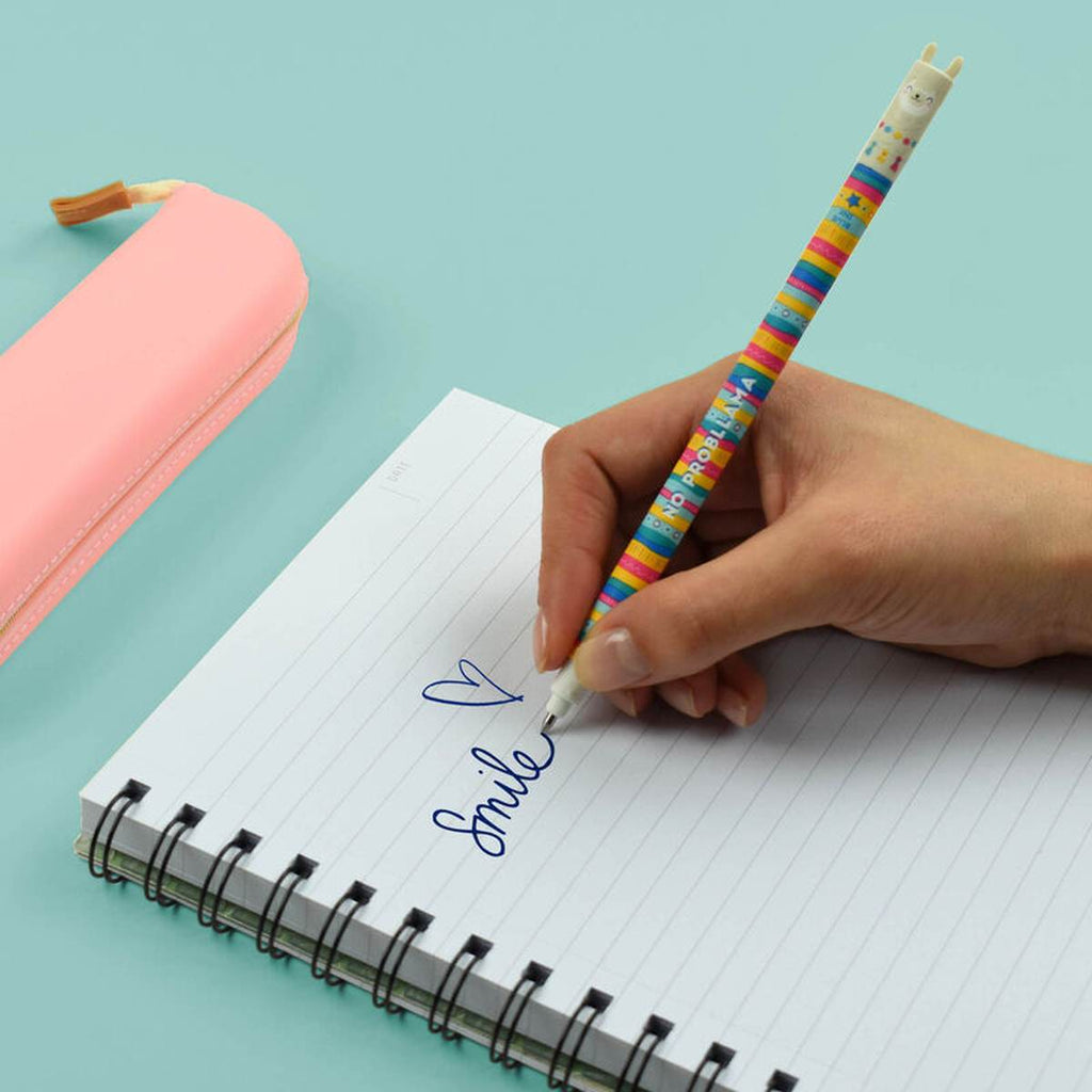 A hand writing on a lined notebook with a slender colourful striped pen with a smiling llama face on the cap on a turquoise desk.