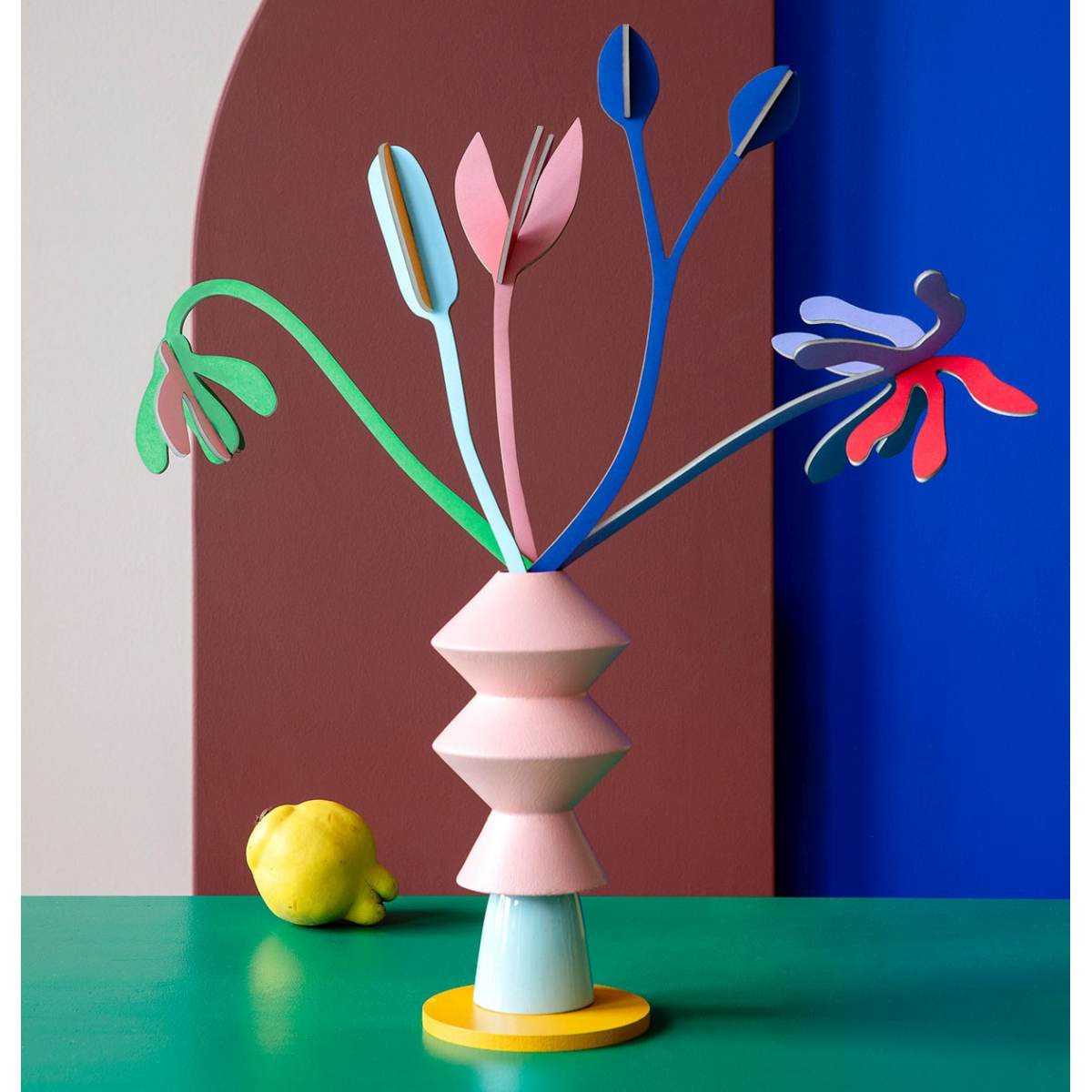 Contained inside a pale pink geometric vase are five playful flower stems that are made out of cardboard and a lemon sitting on the green table in the background.