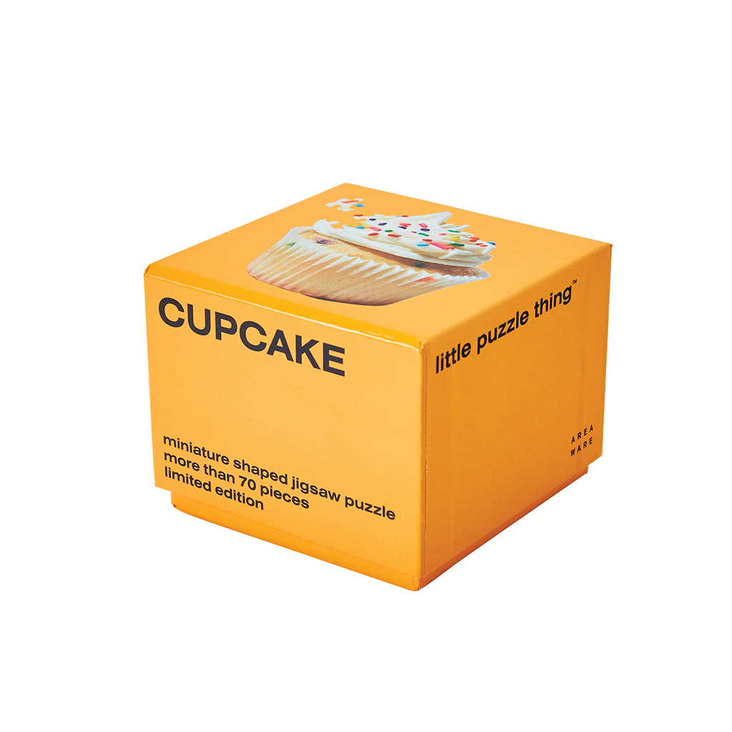 An orange packaging cube has 'cupcake' capitalised in black on one side with 'miniature shaped jigsaw puzzle more than 70 pieces limited edition' at the bottom. The top side has an image of the completed cupcake puzzle with one piece askew. 