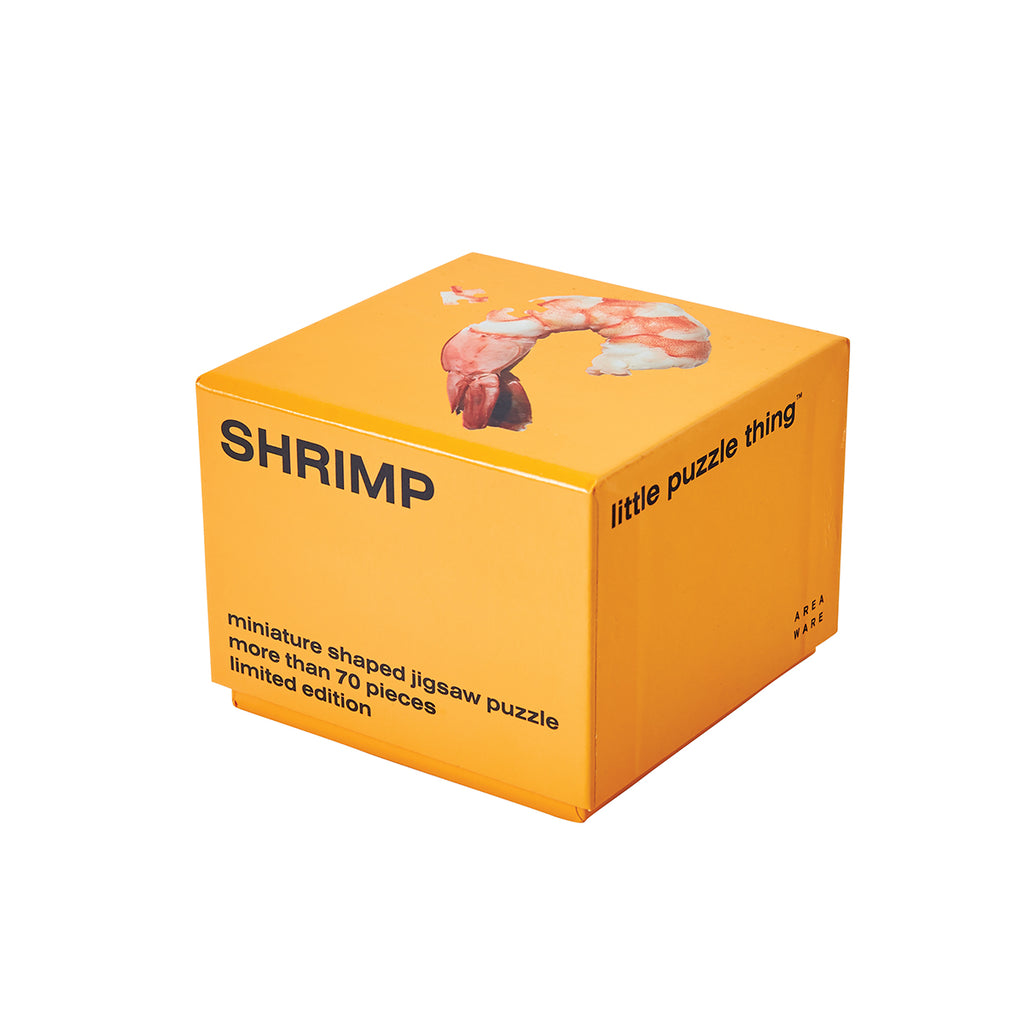 An orange packaging cube has 'shrimp' capitalised in black on one side with 'miniature shaped jigsaw puzzle more than 70 pieces limited edition' at the bottom. The top side has an image of the completed prawn puzzle with one piece ajarred. 