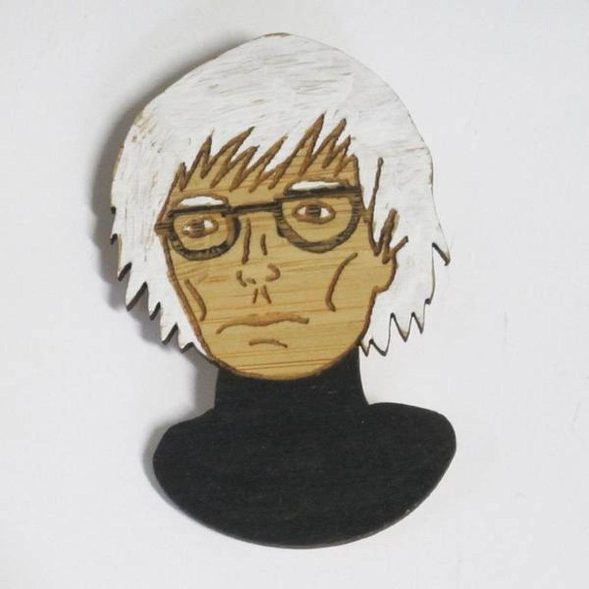 A brooch featuring a portrait of artist Andy Warhol. He is shown wearing a black turtleneck and his iconic glasses.Made from bamboo wood and hand painted.