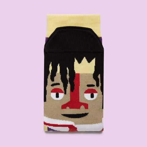A sock with a yellow band is folded on a lavender background shows the toe end with Jean-Michel Basquiat's face and iconic black hair.