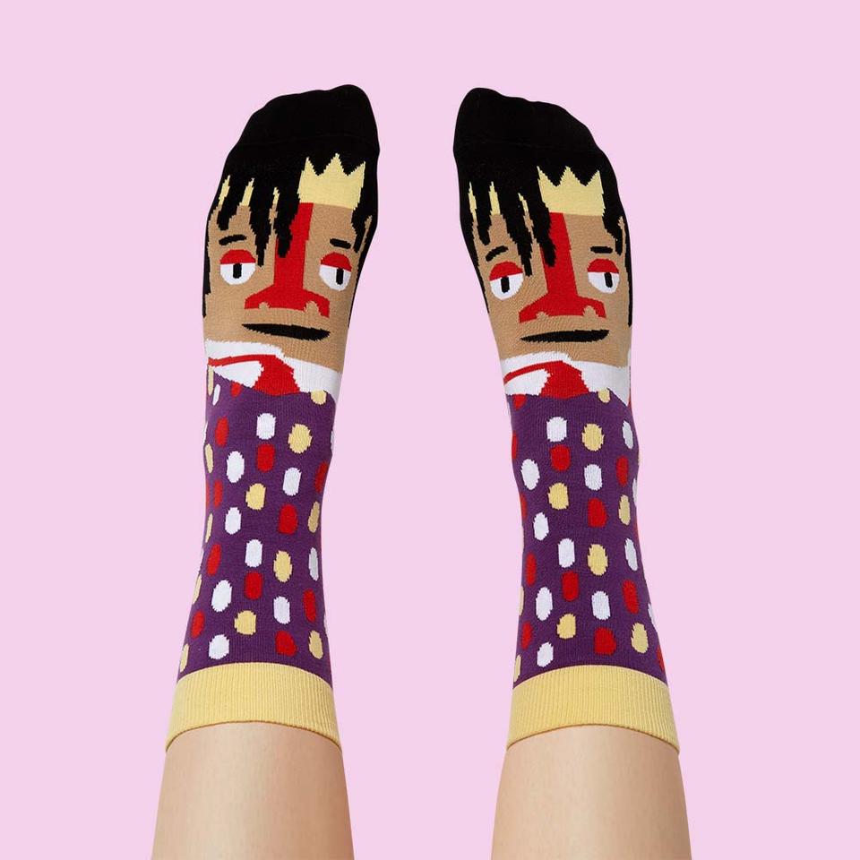 Against a lilac background are a pair of feet wearing purple socks with red, white, yellow polka dots. At the toe end is Jean-Michel Basquiat's face and iconic black hair.