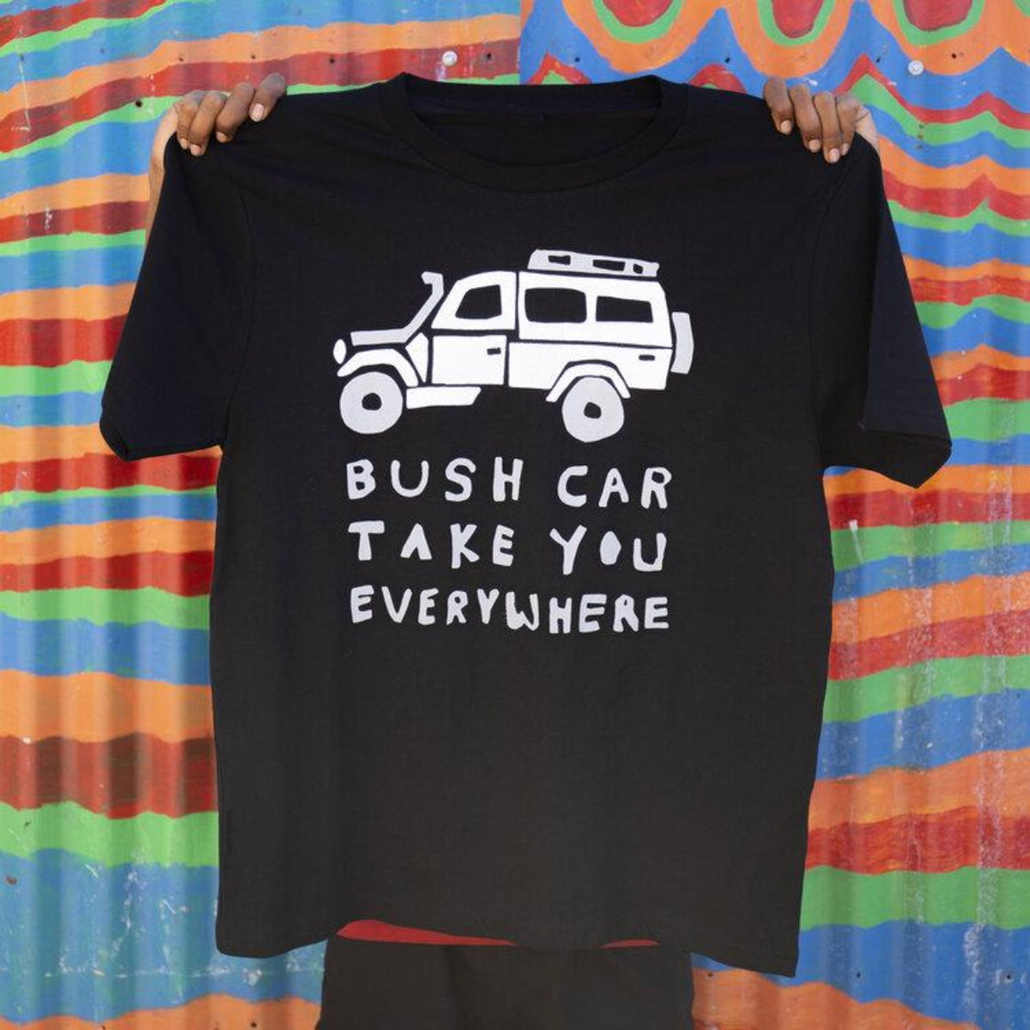 A person displays a black tshirt, holding it up in front of them b the shoulders. An image of a 4WD and text in white features on the black t-shirt. The text reads "Bush Car Take you everywhere"