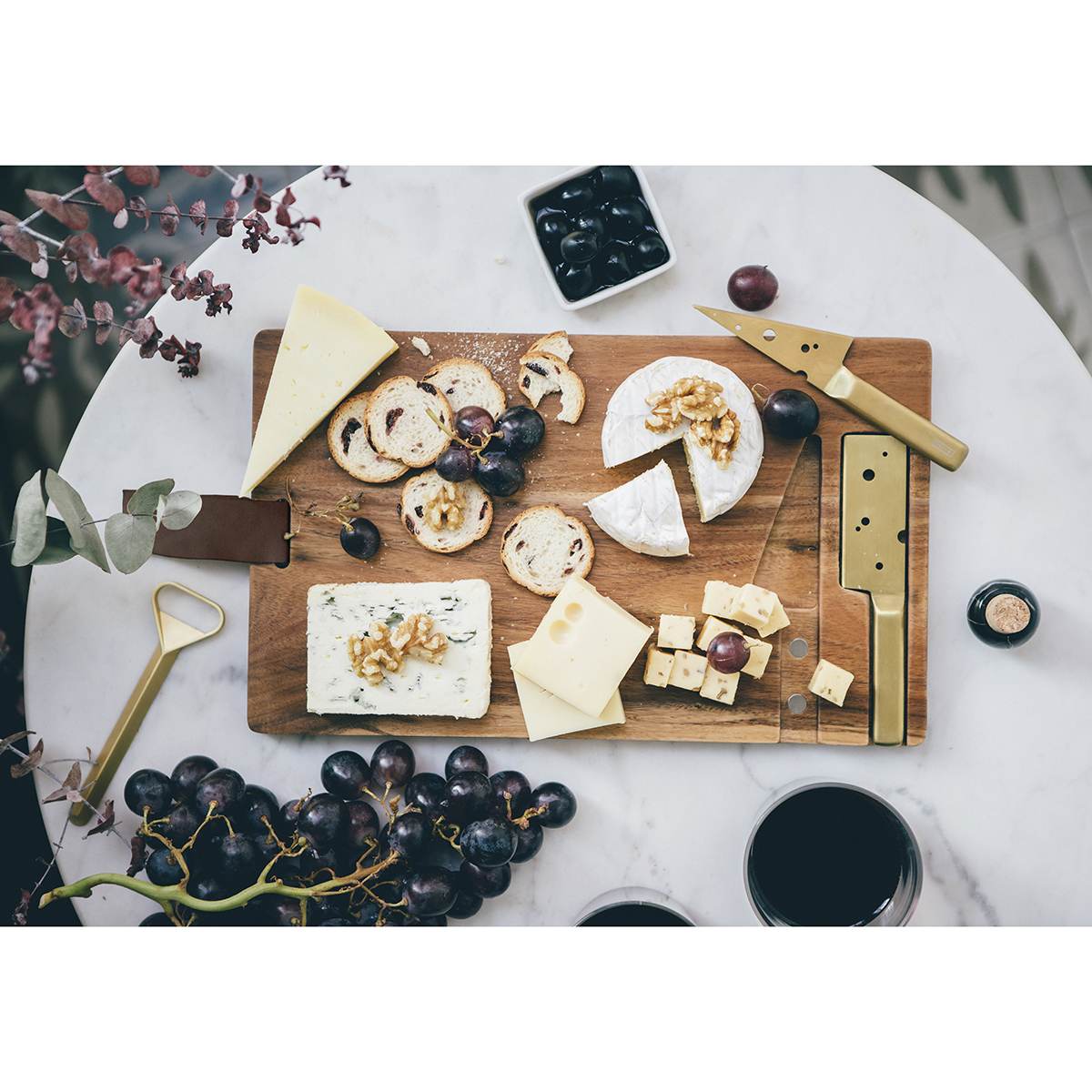 The acacia wood cheeseboard covered with assorted cheeses and crackers that are sliced with the gold cheese holder knife. 