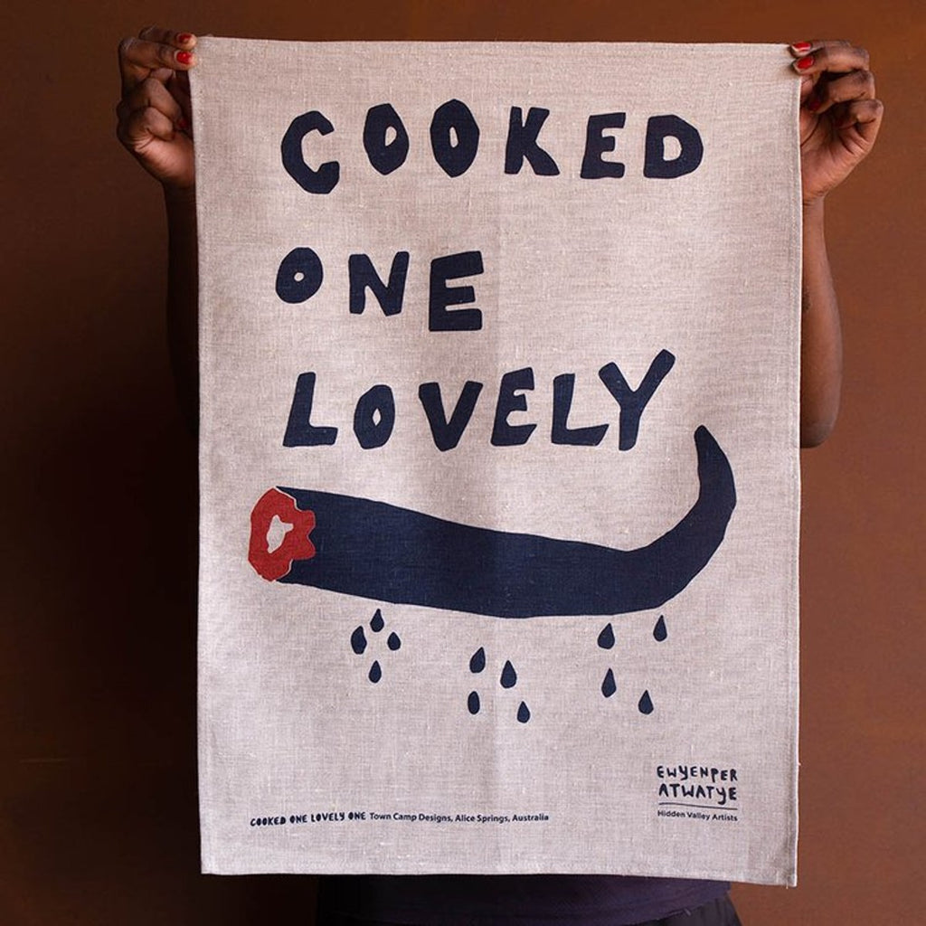 A Natural Linen Tea Towel with a black and red screenprinted work depicting a cooked kangaroo tail. The word 'COOKED ONE LOVELY' appears in bold.
