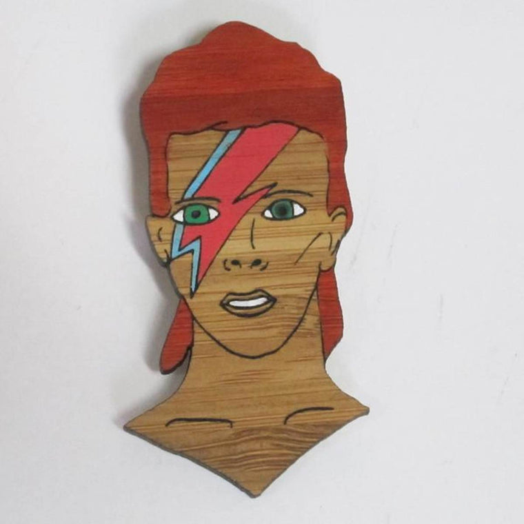 A brooch featuring a portrait of singer songwriter David Bowie. He is shown from the neck up, with his iconic red hair and a blue and red lightning bold painted across his face..Made from bamboo wood and hand painted.