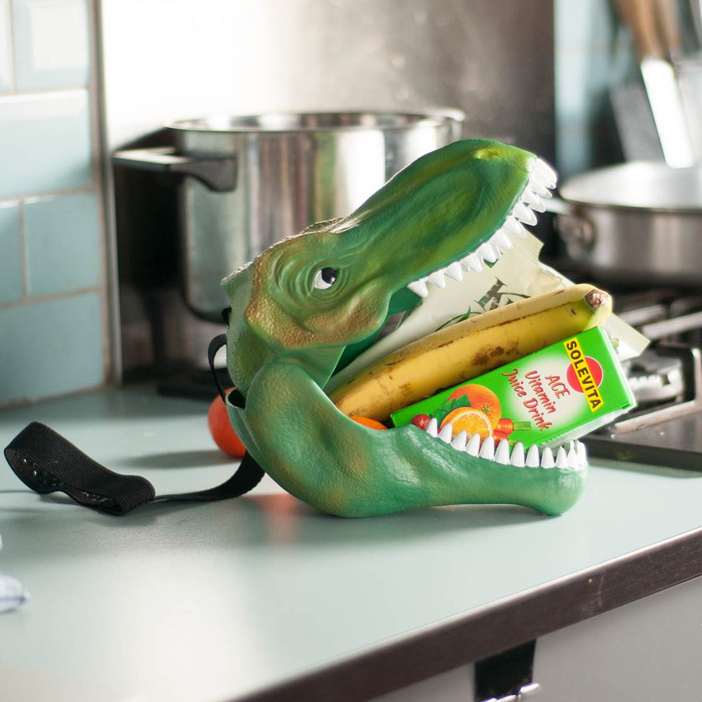A green Tyrannosaur Rex head containing a banana, white packet and juice box in its unhinged jaw sitting on a kitchen bench. 
