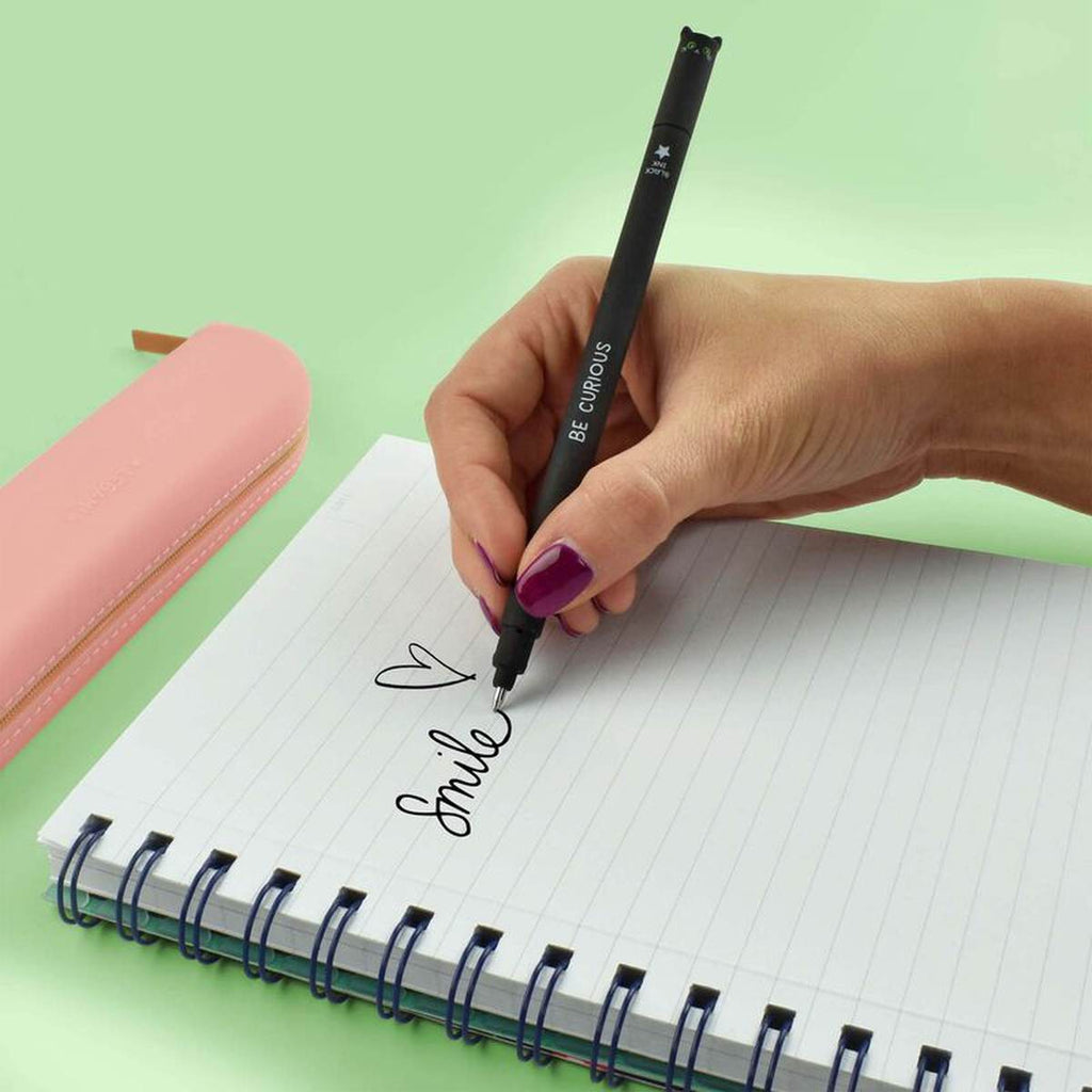 A hand writing on a lined notebook uses a slender black pen with a pig face on the cap on a green desk.