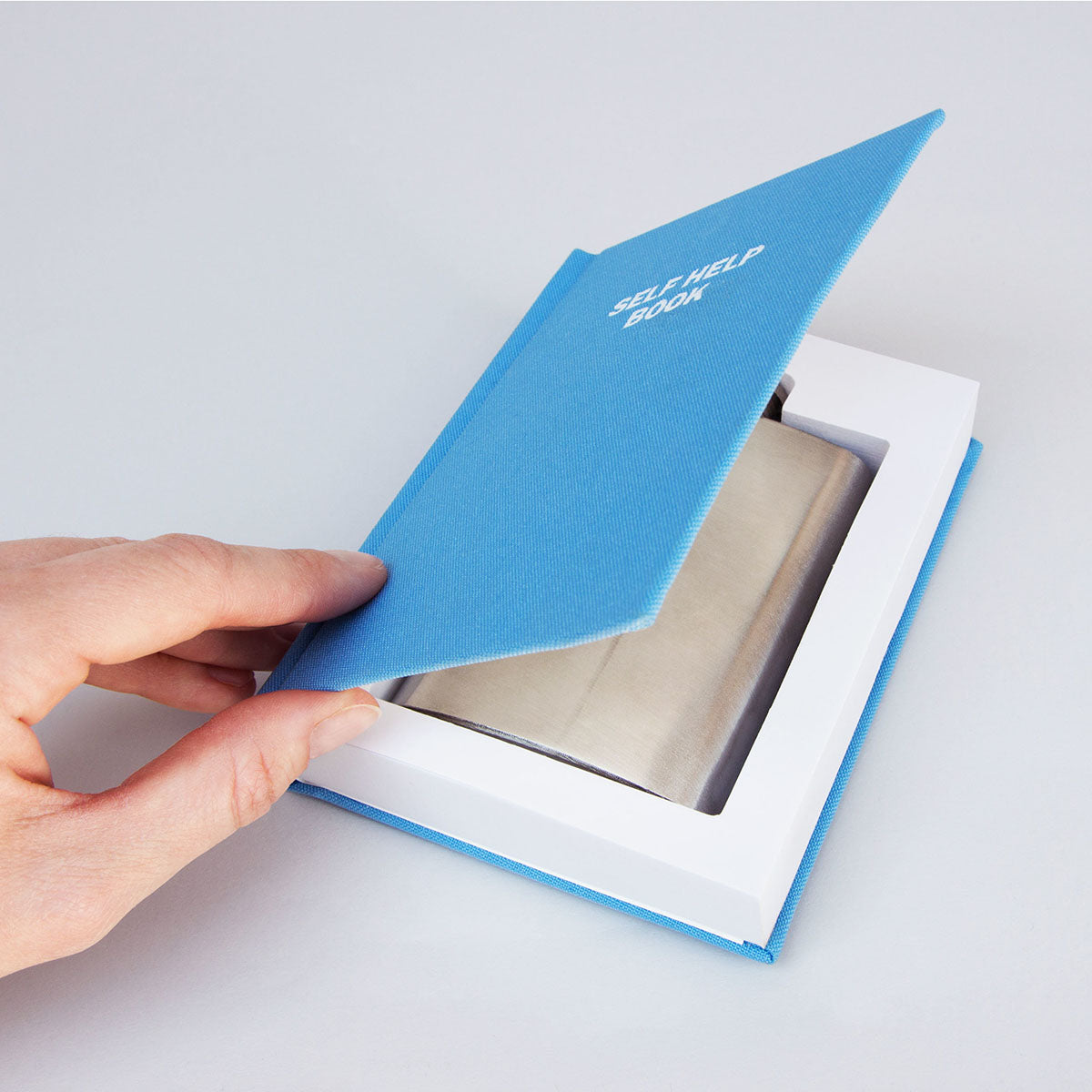 A hand opening the blue cover of the "self help book" reveals a silver drink flask inside the white pages.  