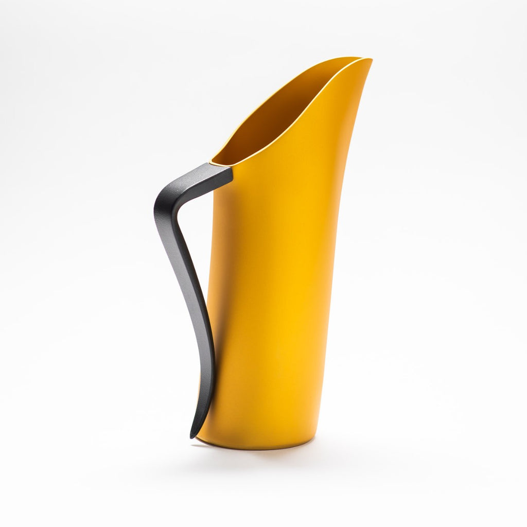 A yellow matte jug with its slender black handle facing towards the front, exposing the sculptural opening of the jug.  