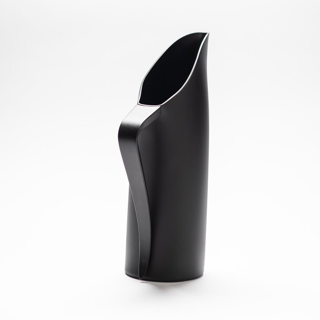 A black matte jug with its slender black handle facing towards the front, exposing the sculptural opening of the jug.  