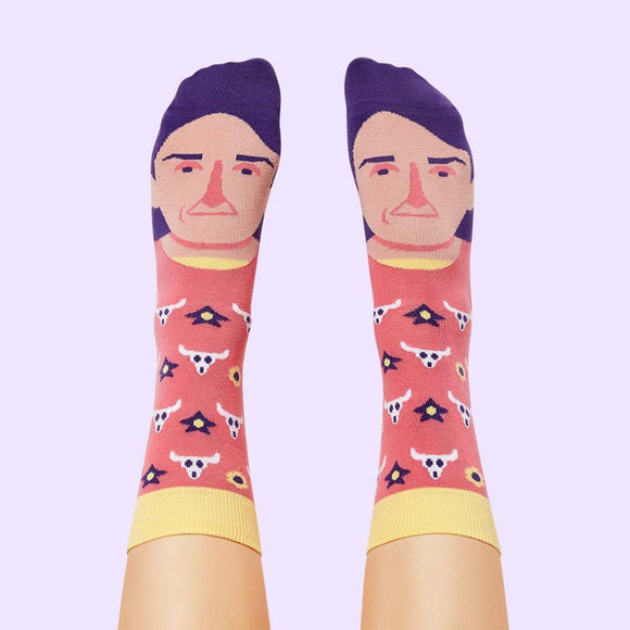 A pink and yellow sock folded on a lavender background, showing the toe end with Georgia O'Keefe's face and purple hair. 