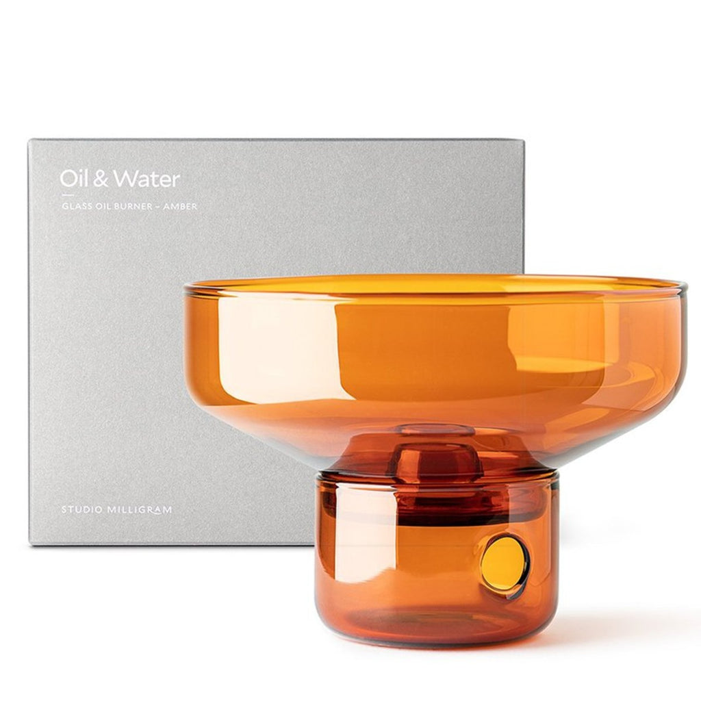 In front of the grey cube packaging box with the text “Oil & Water” on it is a amber glass oil burner. The top is conical shape that sits on top of short cylindrical glass with a round hole on the side.