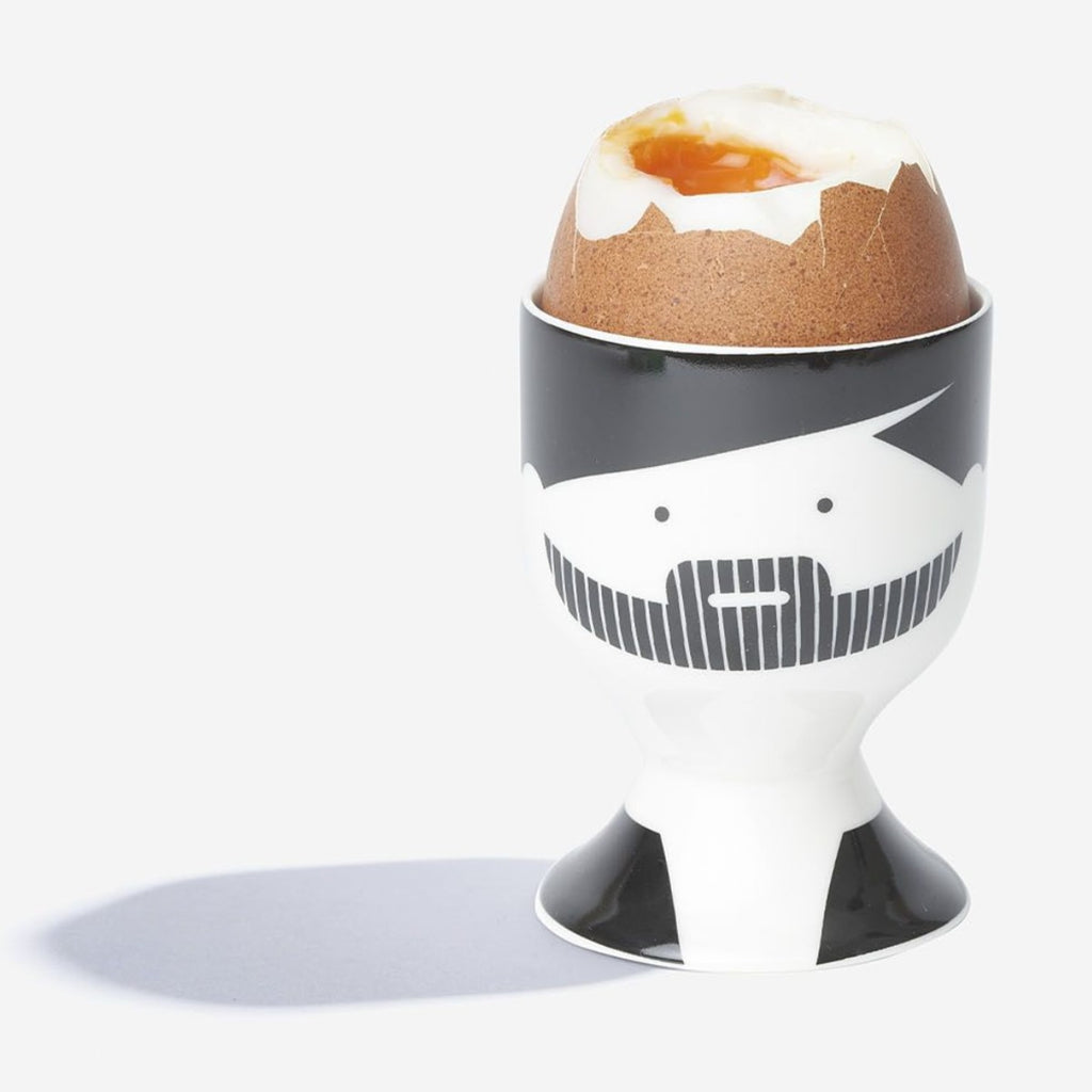 A white porcelain egg cup with Maimonides’ facial features designed in black contains a cracked boiled egg. 