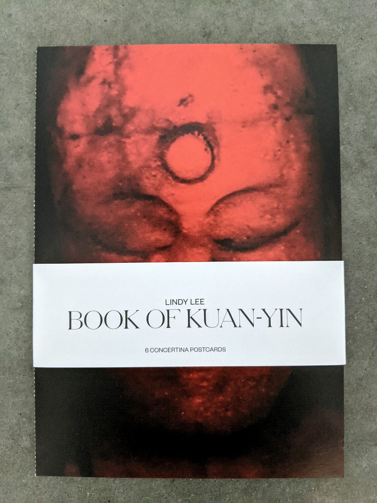 Laying flat on the concrete floor is a postcard with a black and red print of Kuan-Yin bounded by the white band with the text "LINDY LEE BOOK OF KUAN-YIN". 