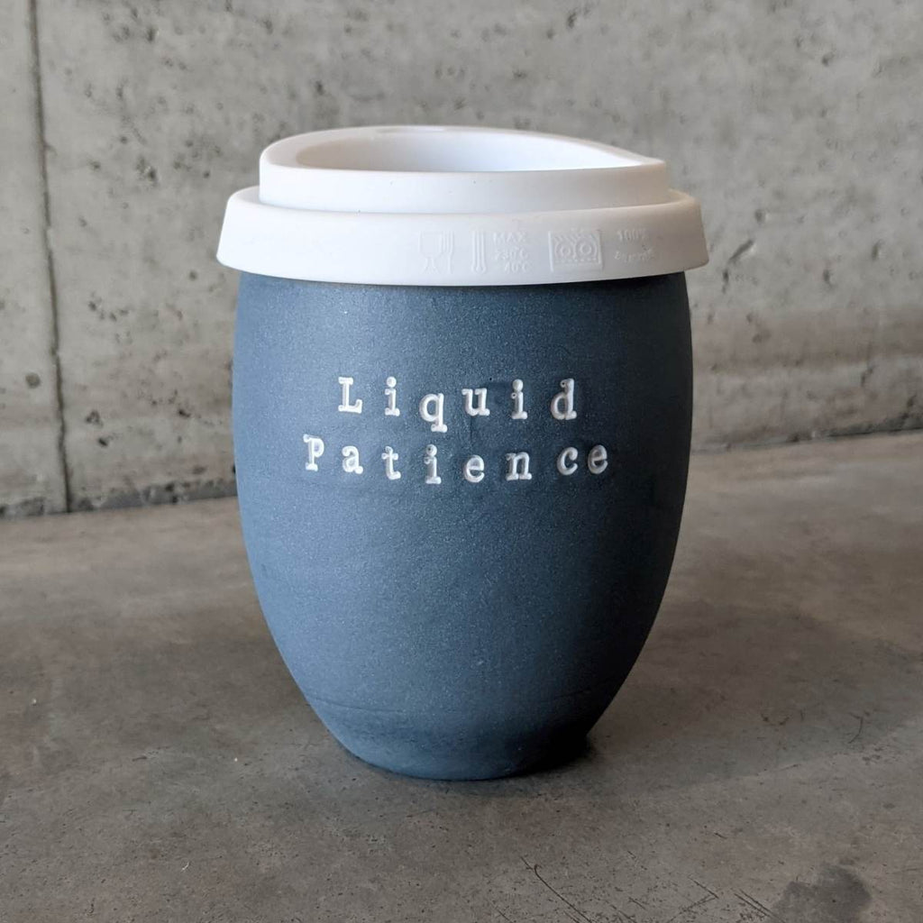 An egg-shaped cup with a flat base made of navy matte ceramic has a 'Liquid Patience' in white typewriter font embossed on the mug and a white sippy lid, like takeaway coffee lids, made of silicone on the top. 