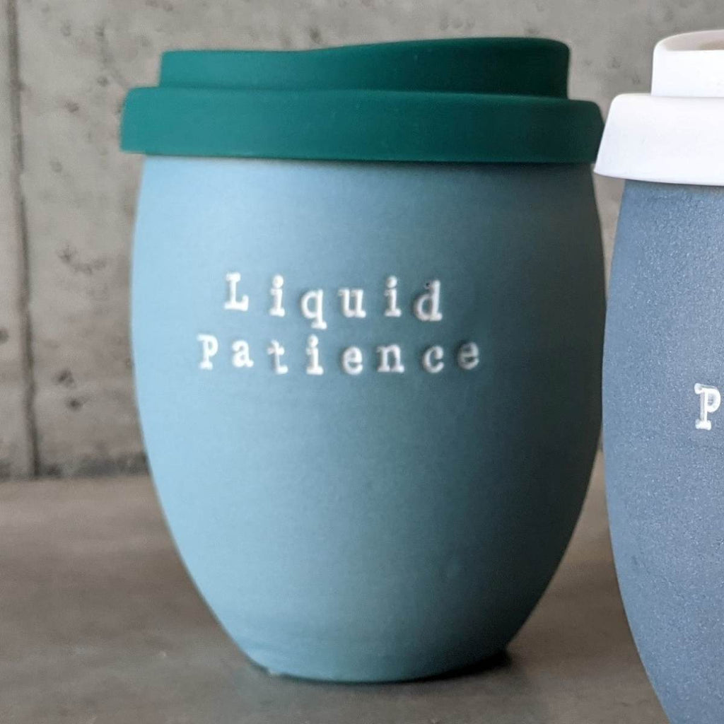 An egg-shaped cup with a flat base made of air force blue matte ceramic has a 'liquid patience' in white typewriter font embossed on the mug and a teal sippy lid, like takeaway coffee lids, made of silicone on the top. 