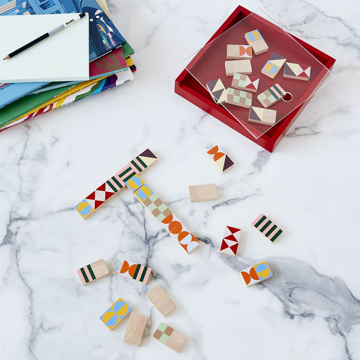 An opened red box of dominoes with a clear lid sits next to a game of dominoes playfully arranged on the marble table. 