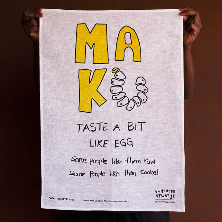 A white linen tea towel with a handwritten design of 'MAK' in bold yellow capitalised font with a row of witchetty grub stylised into the U letter. A blackhandwritten text underneath states 'TASTE A BIT LIKE EGG' capitalised while 'some people like them raw' and 'some people like them cooked' in lowercase.
