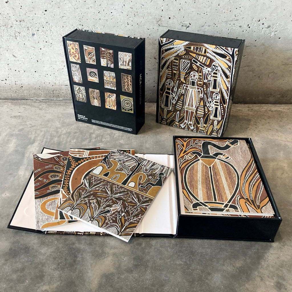 The front and back of a card set containging 12 cards printed with the ochre on bark works by David Malangi Daymirringu. In the foreground one card set is open displaying four card designs.