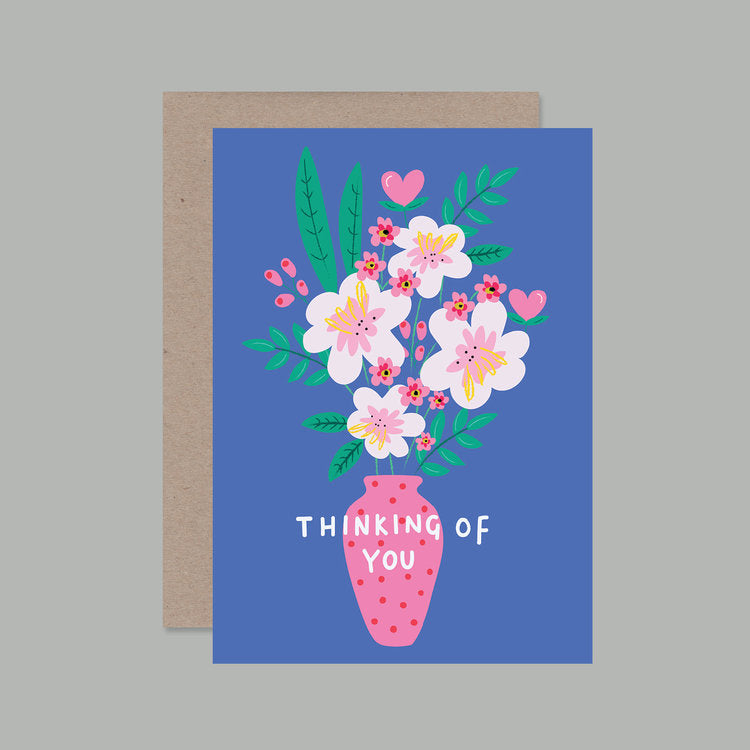 A royal blue greeting card has "thinking of you" in white over a graphic of a pink polka-dot vase holding a bouquet of assorted pink flowers, greenery and pink hearts. 