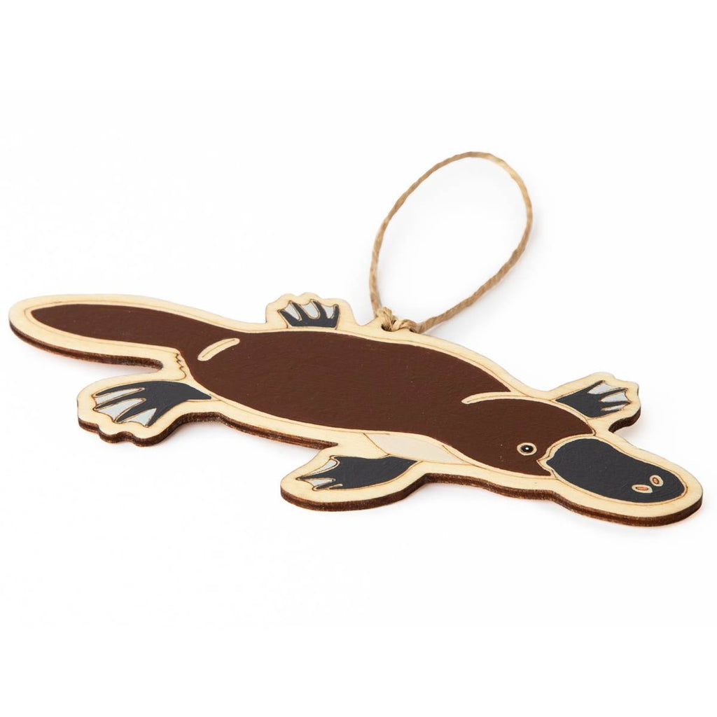 Laying flat at an angle on a white surface is a hanging ornament in the form of a Platypus. Laser Etched flat wood is adorned with brown, black and white hand painting. A Jute string is attached