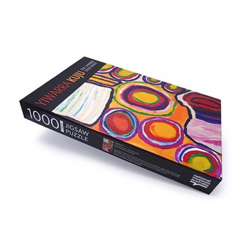 A rectangular packaging box laying flat has 'Yiwarra Kuju' on the top black banner and an image of a painting with a circular pattern in bold, bright colours. The side of the box is black with '1000 pieces jigsaw puzzle' in white. 