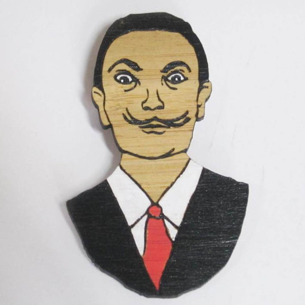 A brooch featuring a portrait of artist Salvador Dali. He is shown wearing a black suit and tie. .Made from bamboo wood and hand painted.