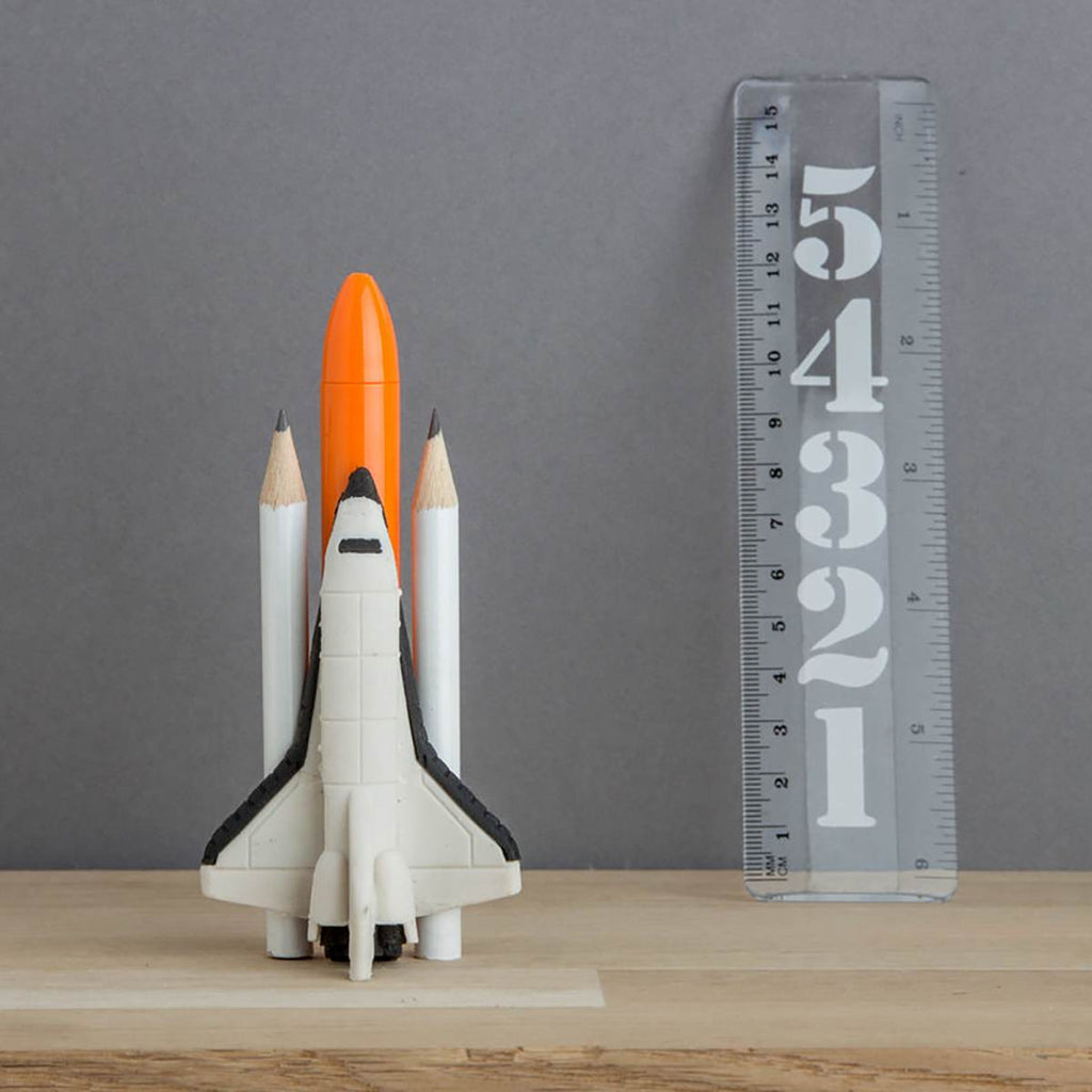 a stylised stationary set compiled to look like a Space Shuttle. Shown is a Space Shuttle eraser, a orange 'rocket' pen, two white pencil 'boosters' and a clear ruler printed with a countdown from 5 to 1.