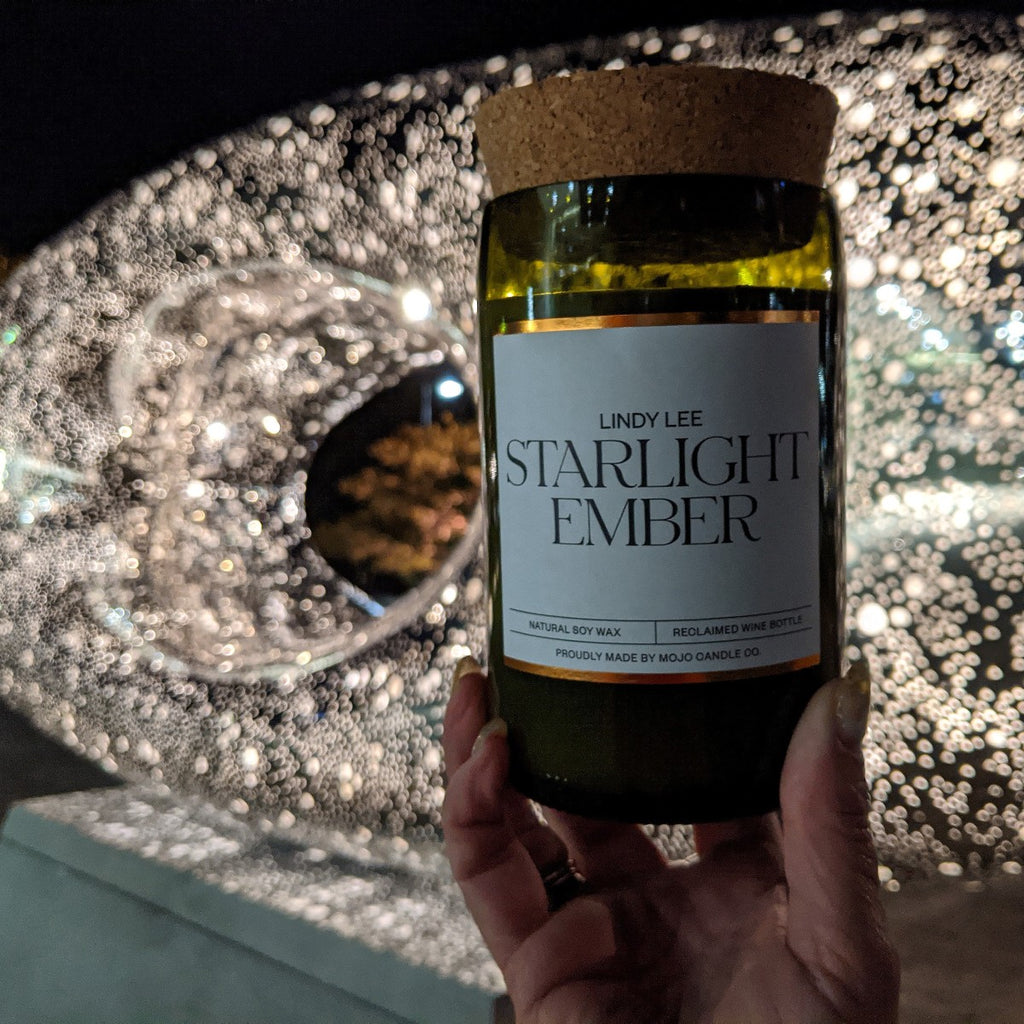 A candle with the label "Starlight Ember" is held by a hand in front of the sculpture 'Secret World of a Starlight Ember'. The sculpture is lit up against a dark night sky.
