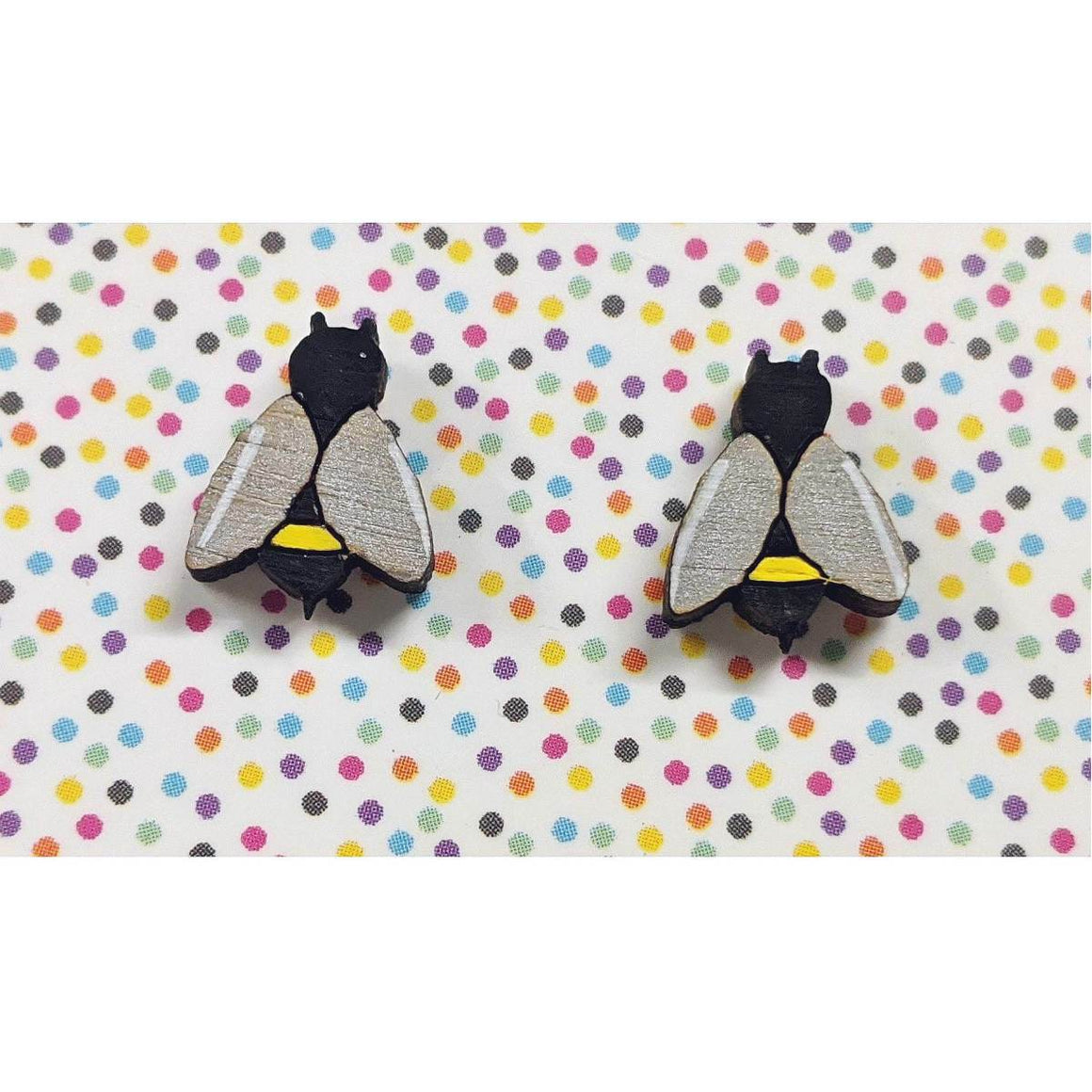 A pair of intricately hand coloured studs depicting honey bees. In yellow and black stripes, their wings are painted in a lustrous silver. Shown on a rainbow polka dot background.