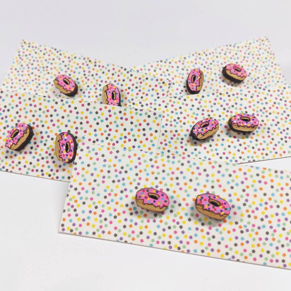 A pair of intricately hand coloured studs depicting pink iced donuts. The pink icing feature rainbow sprinkles. Shown on a rainbow polka dot background.