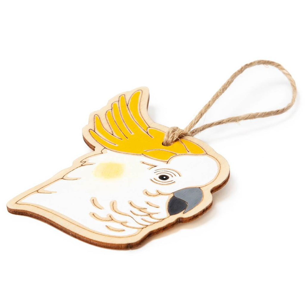 Laying flat at an angle on a white surface is a hanging ornament in the form of a Sulphur-Crested Cockatoo. Laser Etched flat wood is adorned with white, grey, black and yellow hand painting. A Jute string is attached