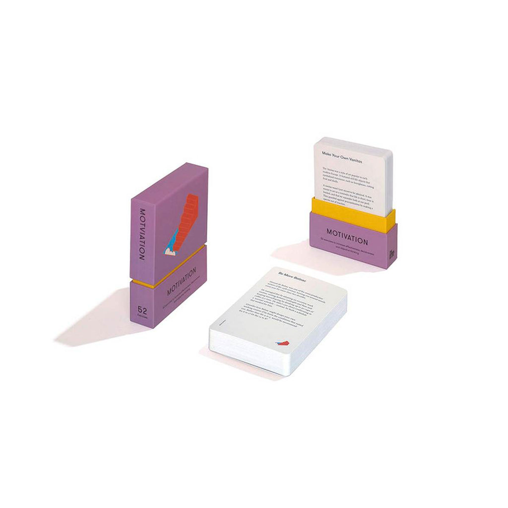 A purple box with a tangerine graphic on the top centre faces the long side of the white cards piled in the centre. Facing the top of the card deck is a lidless purple case with 'motivation' in black and a yellow interior holding a stack of cards upright. 