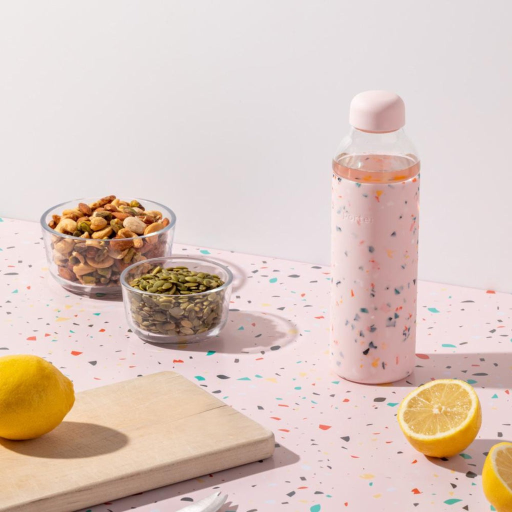 On a pink terrazzo table with colourful speckles is a matching pink terrazzo silicone sleeve on a tall glass bottle among sliced lemons and bowls of nuts and seeds. 
