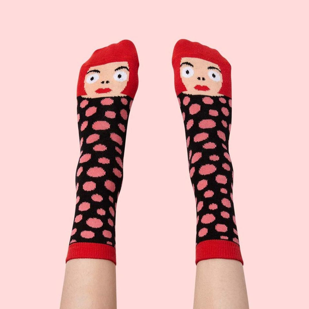 Against the blush pink background are a pair of feet wearing black socks with pink polka dots with Yayoi Kasuma's face and cherry red bob at the toe end.  